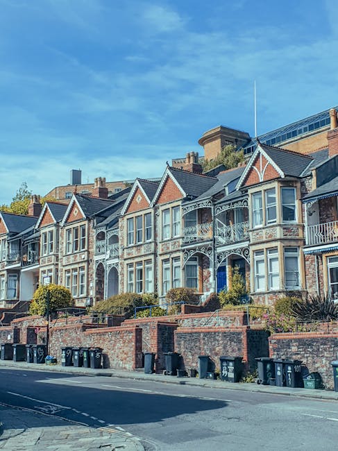 Exterior view of a row of Victorian-style terraced houses with decorated facades, bay windows, and small front gardens bordered by low brick walls and greenery, located along a residential street on a clear day with a bright blue sky. The street features parked trash bins along the pavement, and the image depicts a typical urban neighbourhood scene, relevant to home relocation and house removals services provided by Finsbury Park Man and Van as part of their removals and moving logistics offerings.
