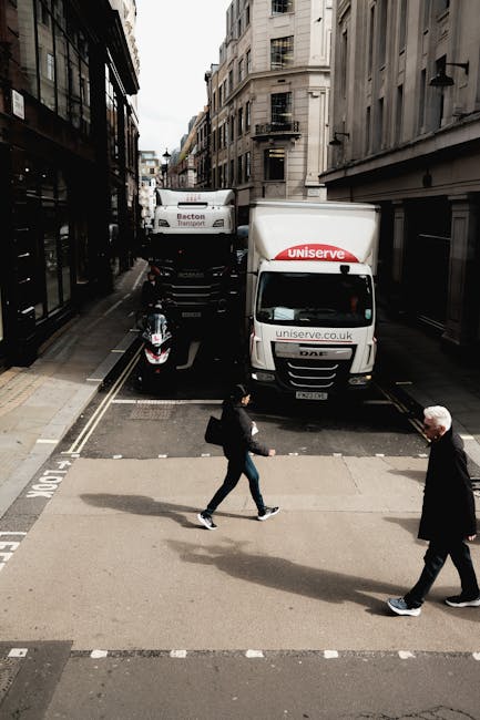 A street scene showing two Finsbury Park Man and Van vehicles engaged in home relocation logistics, with one white moving van parked partially on the pavement at the intersection of narrow urban streets lined with multi-storey buildings. The van displays the website uniserve.co.uk and is associated with the removals service category. Behind the van, another large vehicle is visible, likely used for furniture transport, with a black and white motorbike parked nearby on the road. Two pedestrians, a woman carrying a backpack and a man, are crossing the street on a marked pedestrian crossing. The surroundings are characterized by tall, beige stone buildings with multiple windows and fire escape ladders, under a partly cloudy sky. This setup illustrates the typical loading and unloading process during house removals, with vehicles positioned for efficient loading of packing boxes and furniture within an urban environment, demonstrating the logistical aspects of moving services provided by Finsbury Park Man and Van.