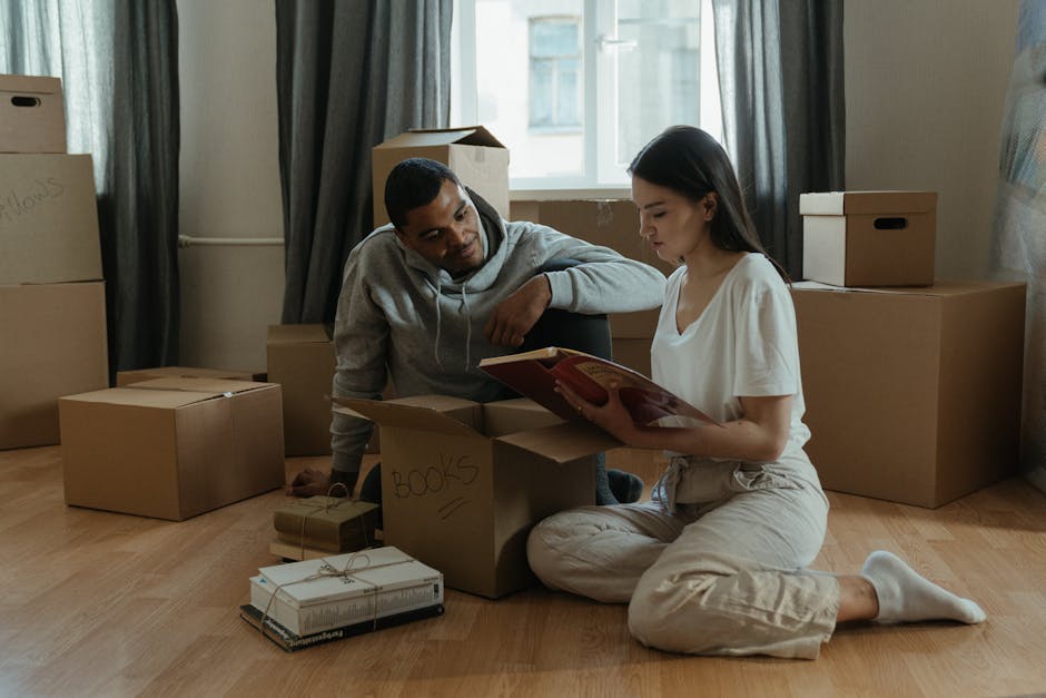 Inside a residential property, two individuals are sitting on a light wood floor surrounded by packed cardboard boxes, some labeled with words like 'books'. The woman, dressed in a white top and beige trousers, is seated cross-legged and reading a book or document, while the man, wearing a grey hoodie, leans on a box and looks on. The scene features natural light coming through a window with open white windowpanes, framed by dark curtains. Various packing materials, including cardboard boxes of different sizes, some with cut-out handles, and a small wrapped package tied with string, are visible. The setting appears to be a home being prepared for a house move, with the packing process in progress. The environment indicates a typical space used for furniture transport and moving preparations, managed by a professional removals service such as Finsbury Park Man and Van.