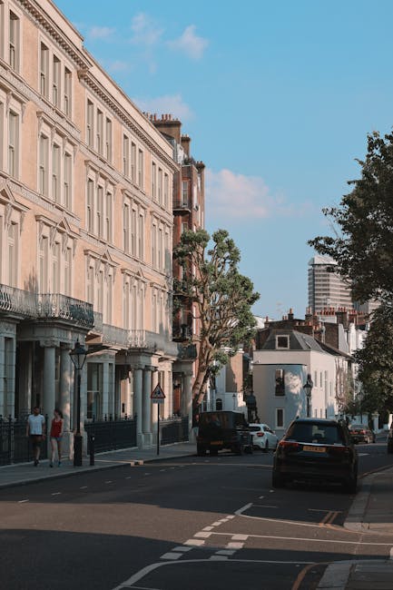 An urban street scene during daytime with a row of multi-storey residential buildings featuring classic architectural details, large windows, and balconies, with some displaying wrought-iron railings. Several trees line the sidewalk, providing greenery, and a few pedestrians are visible walking along the pavement. Parked cars are situated close to the buildings, and a black van is partially visible on the road, suggesting activity related to home relocations or deliveries. The street has marked parking bays and a clear road surface, with a bright blue sky overhead. The scene reflects a typical residential area potentially suitable for house removals and furniture transport services, as provided by Finsbury Park Man and Van, supporting efficient packing and moving logistics in Finsbury Park N4.