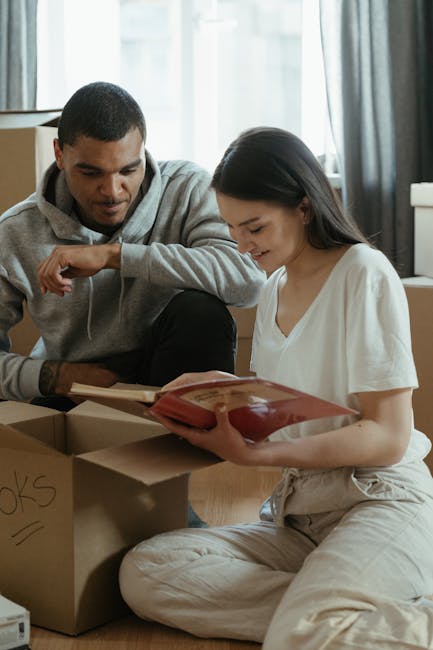 Inside a residential property, two individuals are sitting on a light wood floor surrounded by packed cardboard boxes, some labeled with words like 'books'. The woman, dressed in a white top and beige trousers, is seated cross-legged and reading a book or document, while the man, wearing a grey hoodie, leans on a box and looks on. The scene features natural light coming through a window with open white windowpanes, framed by dark curtains. Various packing materials, including cardboard boxes of different sizes, some with cut-out handles, and a small wrapped package tied with string, are visible. The setting appears to be a home being prepared for a house move, with the packing process in progress. The environment indicates a typical space used for furniture transport and moving preparations, managed by a professional removals service such as Finsbury Park Man and Van.