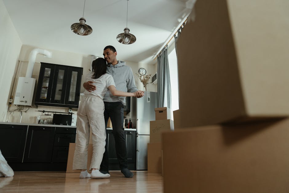 Inside a residential kitchen area, a man and woman are embracing in a moment of celebration or gratitude during a home relocation process. The man, dressed in a light grey hoodie and dark trousers, has his arms around the woman, who is wearing white trousers and a white top, with her arms draped around his neck. The kitchen features black cabinetry with glass-fronted upper cabinets, a white wall-mounted boiler on the left, and a clock with a floral arrangement on the wall behind them. Natural light enters through a window on the right, which is partially covered by a blue curtain. Several cardboard moving boxes are stacked on the floor to the right, indicating packing and moving activities. The scene appears to be part of a furniture transport or packing process, with focus on the emotional moment during the move, supported by the professional services of Finsbury Park Man and Van as they assist with house removals and home relocations.