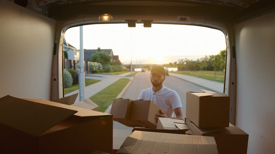 A man wearing a white polo shirt and glasses is loading cardboard moving boxes into the back of a white van during a house removal process. The van's rear doors are open, revealing several stacked cardboard boxes of various sizes, some sealed with packing tape and others with flaps open. Outside, a residential street at sunset is visible, with a sidewalk, a streetlamp, bushes, and houses in the distance. The scene indicates a packing and moving activity as part of a home relocation, with the man carefully placing boxes into the vehicle, supported by Finsbury Park Man and Van's professional removals services.