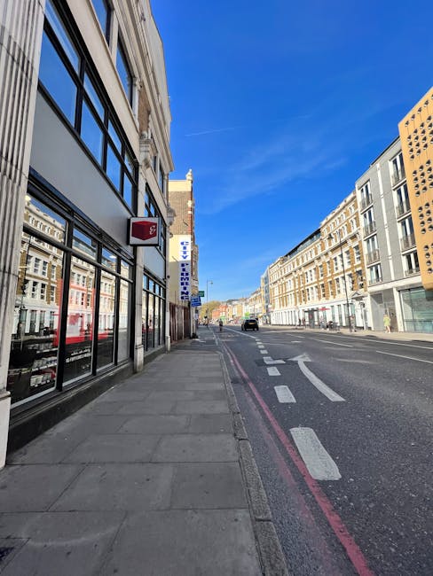 Photograph of a quiet city street in Finsbury Park with a clear blue sky overhead. The left side features a commercial building with large glass windows and a red and white sign attached to the facade. The sidewalk runs alongside the building, with a few small objects visible near the window. On the right side, there are multi-storey residential or commercial buildings with varied architectural styles, some with balconies and large windows. The street has a dedicated bus lane marked by red lines, and a black car is visible in the distance, driving away. The scene shows no active moving or loading activity, but the spacious street and sidewalk suggest conditions suitable for furniture transport or home relocation services, as provided by Finsbury Park Man and Van.