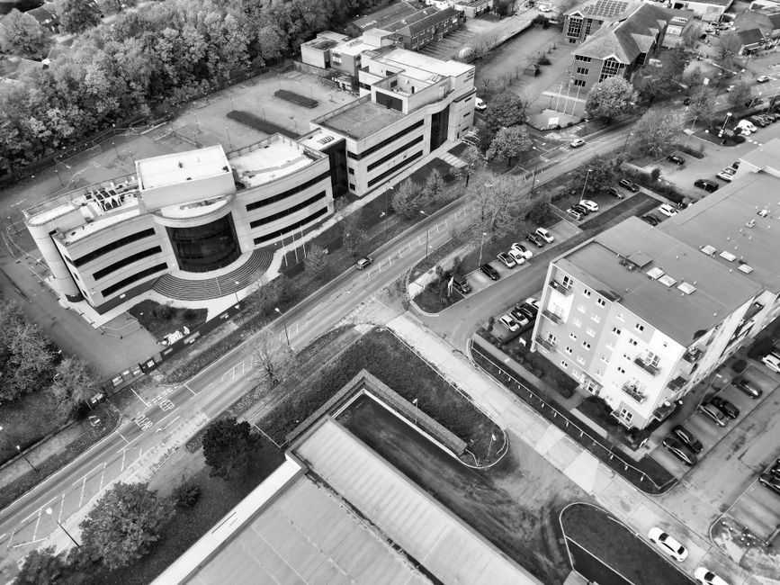A black-and-white aerial photograph showing a modern, curved office building with multiple floors and large glass windows situated next to a parking lot filled with cars. Adjacent to the office, a smaller residential building with several balconies is visible, along with surrounding streets, trees, and additional parking areas. The scene captures the loading area and surrounding pavement where furniture, boxes, and equipment might be prepared for home relocation or furniture transport, with some vehicles parked nearby, likely used by Finsbury Park Man and Van for their removals service tailored to businesses and households in Finsbury Park Blackstock Road.