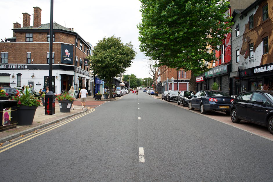 Photograph of a quiet city street in Finsbury Park with a clear blue sky overhead. The left side features a commercial building with large glass windows and a red and white sign attached to the facade. The sidewalk runs alongside the building, with a few small objects visible near the window. On the right side, there are multi-storey residential or commercial buildings with varied architectural styles, some with balconies and large windows. The street has a dedicated bus lane marked by red lines, and a black car is visible in the distance, driving away. The scene shows no active moving or loading activity, but the spacious street and sidewalk suggest conditions suitable for furniture transport or home relocation services, as provided by Finsbury Park Man and Van.