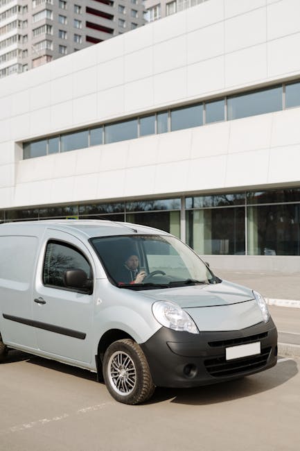 A white commercial van parked on a paved street in front of a modern, curved white building with large glass windows. The van's driver is visible through the windshield, holding the steering wheel, with a neutral background of tall residential or office buildings behind the structure. The van is positioned near the curb, possibly preparing for a home relocation or furniture transport. Inside the van or nearby, there are packing materials such as cardboard boxes, plastic wrapping, and foam padding, indicating ongoing moving or packing activities. The vehicle has black side mirrors, black bumpers, and multi-spoke alloy wheels. The setting appears to be during daytime with natural light illuminating the scene, and the environment is tidy, reflecting a professional move coordinated by Finsbury Park Man and Van as part of their removals service in the Finsbury Park area.